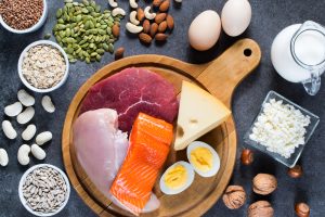 Alimentos ricos em proteína vistos de cima: carne, peixe, laticínios, ovos, trigo, aveia, nozes, feijão, sementes de abóbora e sementes de girassol.