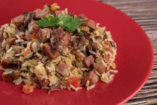 Prato de arroz carreteiro servido em uma travessa vermelha sobre superfície de madeira. O arroz está misturado com pedaços de linguiça, carne bovina em cubos, tomate picado e ervas finas.