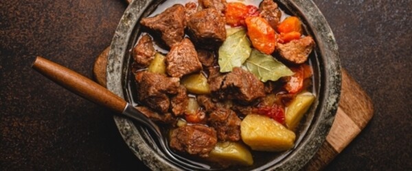 Tigela de ensopado de carne sobre superfície de madeira, com pedaços de carne cozida, cenoura, batata e tomate mergulhados em caldo.