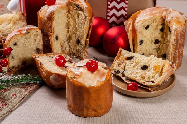 Uma foto de sobremesas de Natal, especificamente mini panetones caseiros com frutas e nozes. A imagem mostra vários panetones em forminhas decoradas com padrões de flores brancas. Cada panetone é coberto com uma camada de glacê branco e decorado com uma cereja vermelha no topo. 