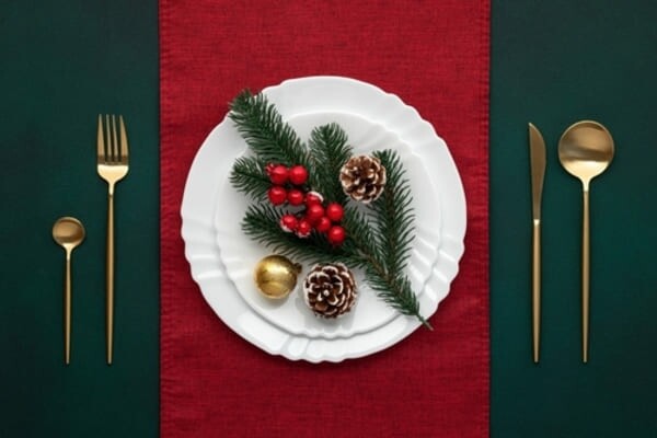 Mesa de Natal elegantemente posta com louças brancas, guardanapos vermelhos, enfeites de pinha e ramagens verdes, taças de cristal e detalhes em dourado.