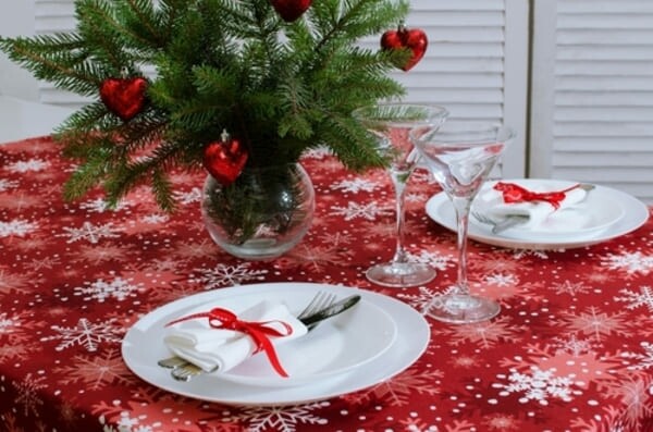 Mesa de jantar decorada para o Natal com louças elegantes, velas acesas e uma toalha temática vermelha com estampas natalinas brancas, criando um ambiente acolhedor e festivo.