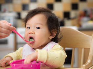 Bebê asiática sorridente sentada em uma cadeirinha de alimentação, experimentando sua primeira comida sólida, usando um babador e em um ambiente de cozinha iluminado e acolhedor.