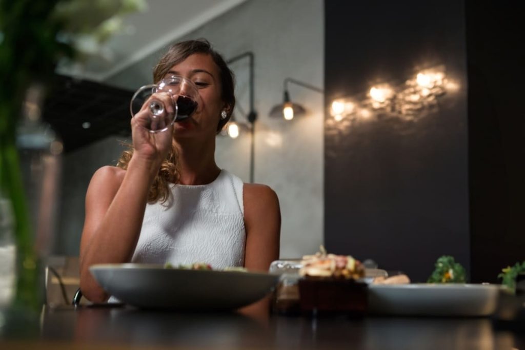 Mulher apreciando uma taça de vinho tinto sentada à mesa de madeira, com foco na atmosfera acolhedora e intimista.