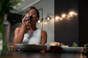 Mulher apreciando uma taça de vinho tinto sentada à mesa de madeira, com foco na atmosfera acolhedora e intimista.