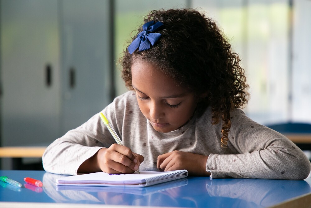 Menina concentrada sentada à mesa da escola, desenhando em um caderno durante a aula.