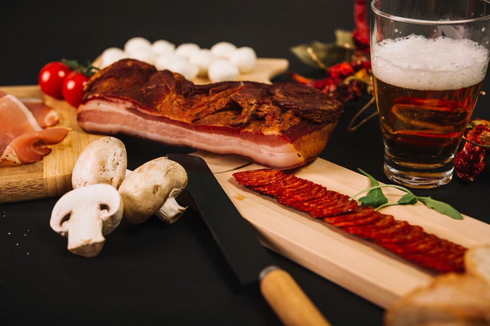 Close-up de ingredientes para preparo de receita, incluindo copo de cerveja e pedaços de carne sobre tábua de madeira.