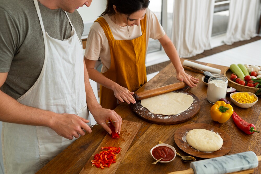 Vista superior de pessoas preparando pizza juntas em casa, com ingredientes e massa sobre a mesa.