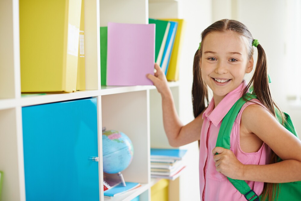 Menina em pé na sala de aula, sorrindo enquanto pega um pequeno caderno da prateleira.